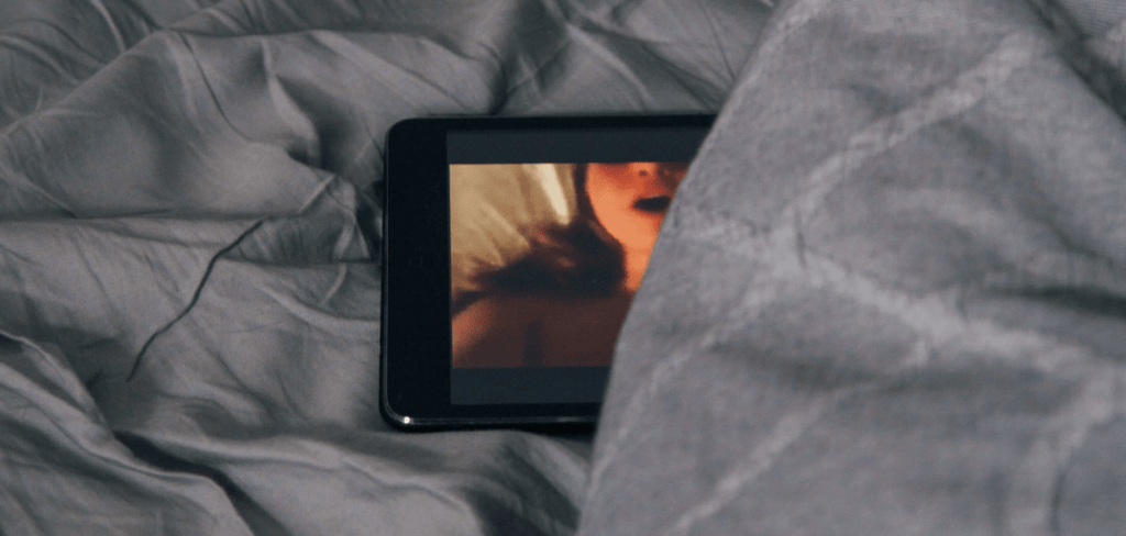 a tablet peeking out of the sheets with porn on the screen
