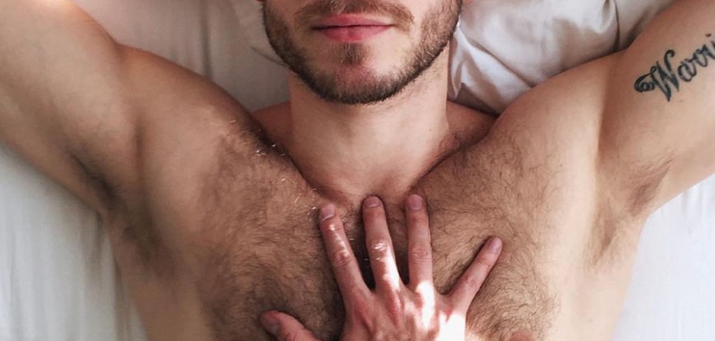 Bare-chested hair man laying in bed with another man's had on his chest