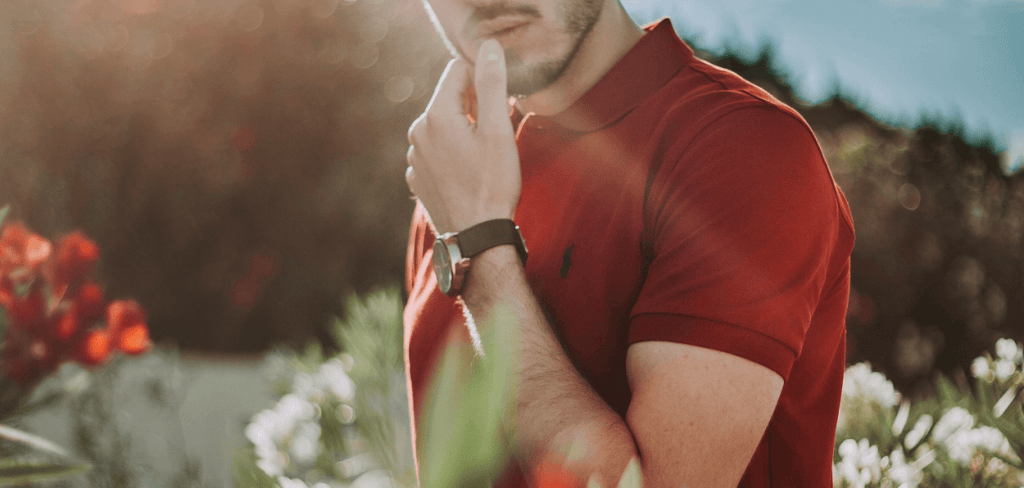 A man posting in a field of flowers wearing a red polo shirt with thumb touching his lips