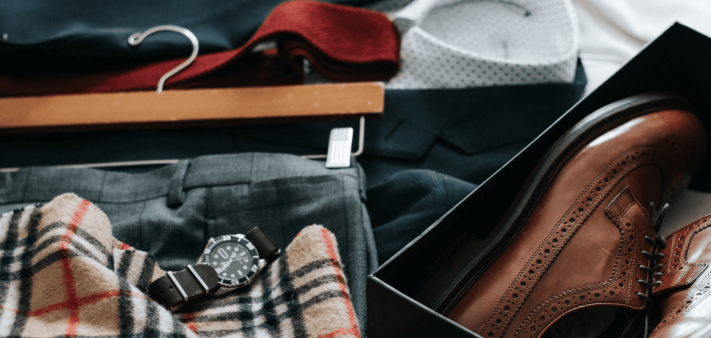 a pair of brown dress shoes in a box with a pair of slacks on a hanger, a watch and scarf on a bed