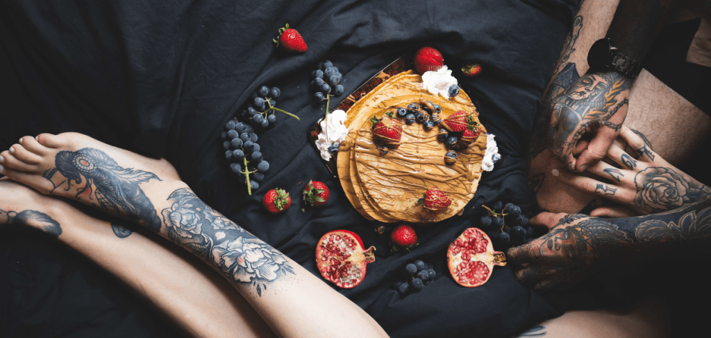 A tattooed couple laying on black sheets eating a bowl of fruit for breakfast