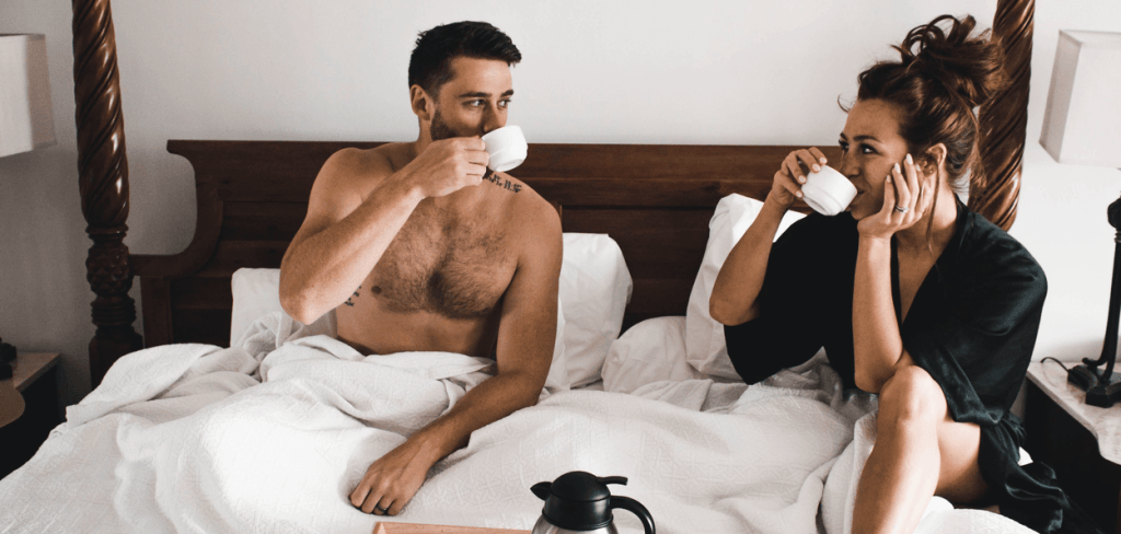 A sexy couple laying in bed laughing and having coffee