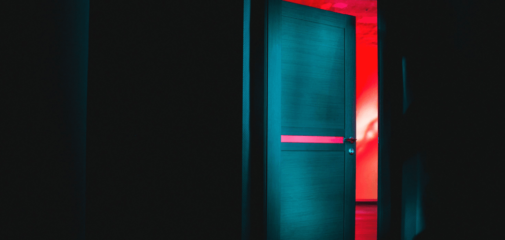 A photo of a door half open in the dark with light with a room wall on the other side