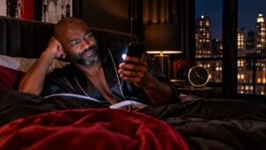 Man in a black silk robe in bed late at night, squinting at a text on his phone, with a clock and city lights behind him