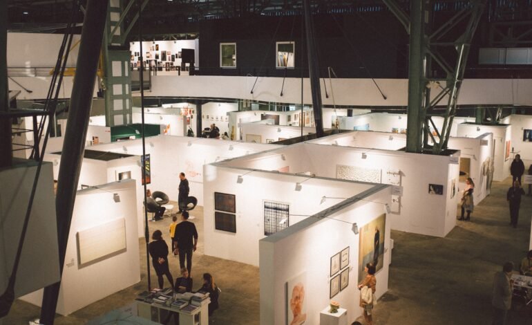 Visitors browsing artwork in exhibition booths inside a large industrial hall.
